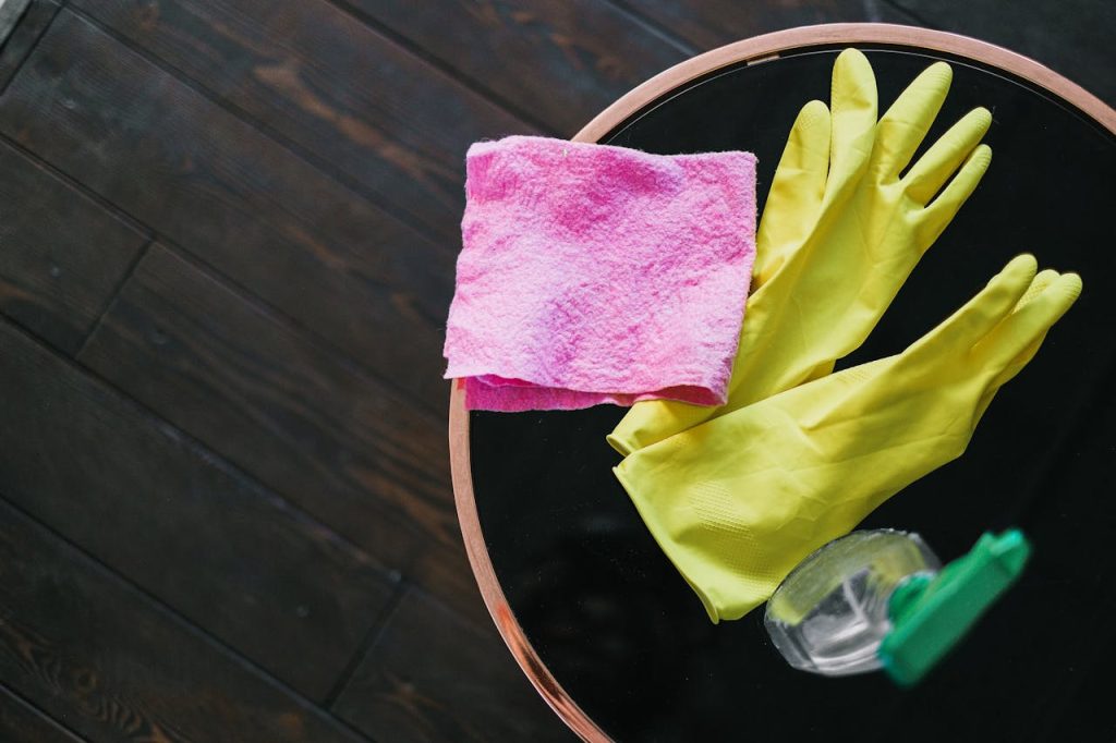Pink microfibre cloth, yellow rubber gloves and a spray bottle on a round glass side table with a copper rim