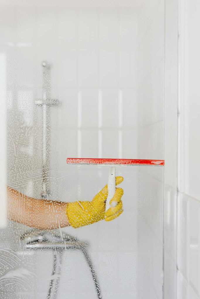 Hand in a yellow rubber glove clearing soap from a glass shower screen with a red squeegee in a white tiled bathroom