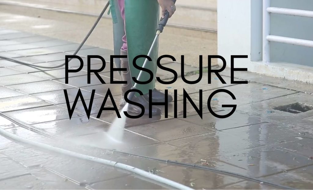Cleaner in a green apron and boots jet washing wet paving slabs outside a building