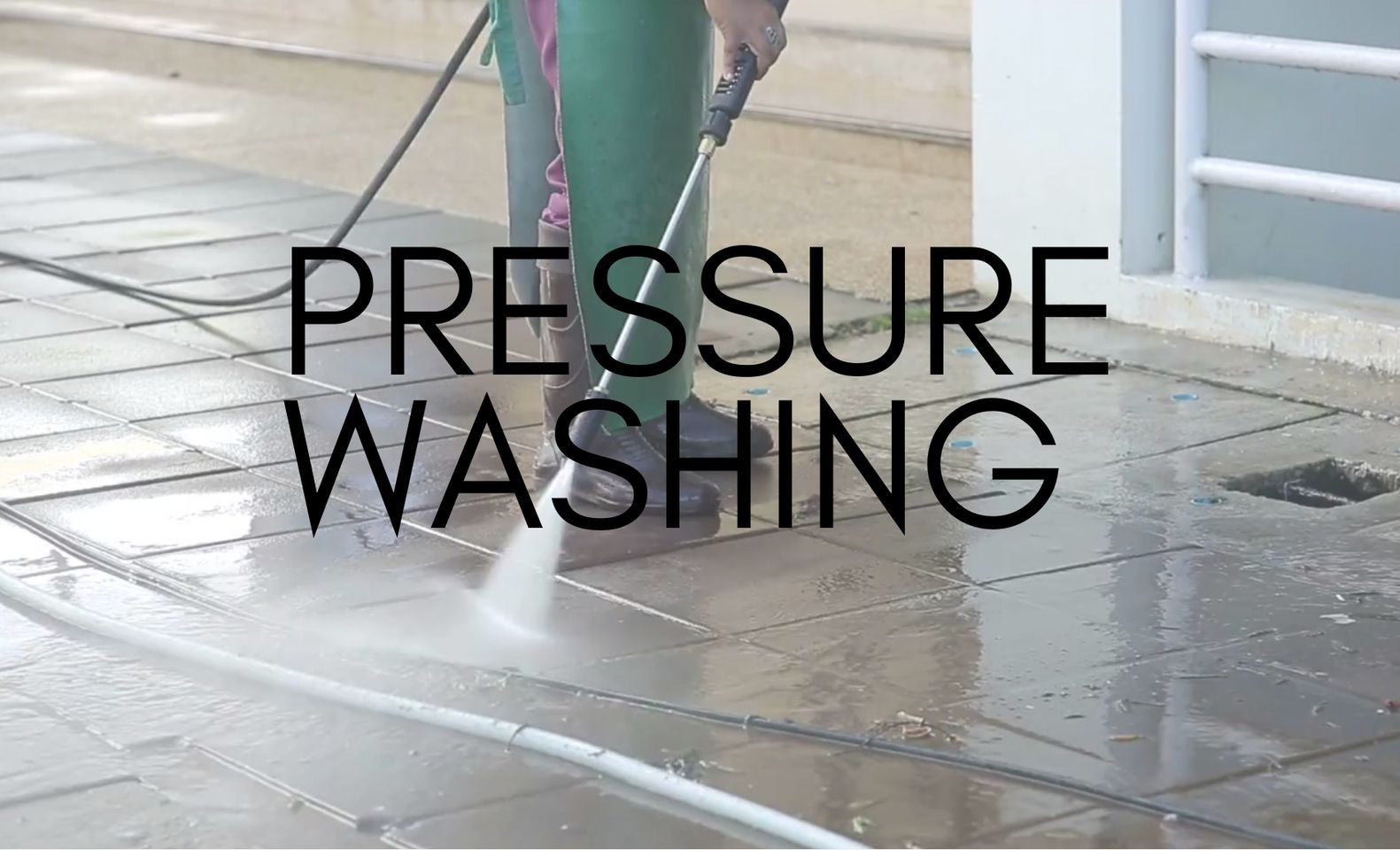 Cleaner in a green apron and boots jet washing wet paving slabs outside a building